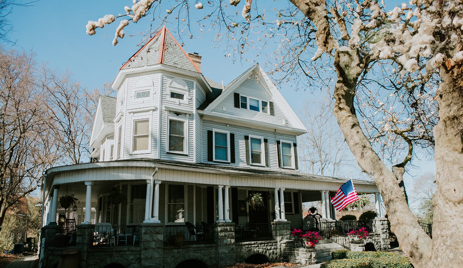 A large, white Victorian-style house with a red roof, featuring a porch and an American flag, surrounded by trees.