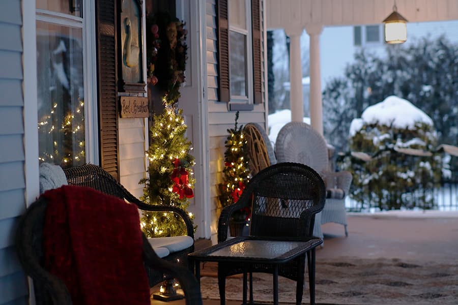 A cozy porch decorated with small Christmas trees and a red blanket, framed by a snowy landscape.