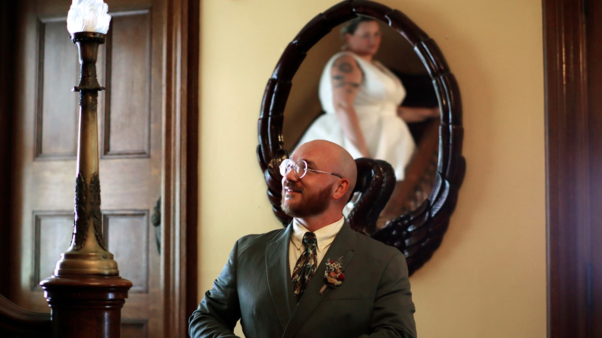 A man in a suit smiles while standing next to a lamp, with a woman in a white dress reflected in a nearby mirror.