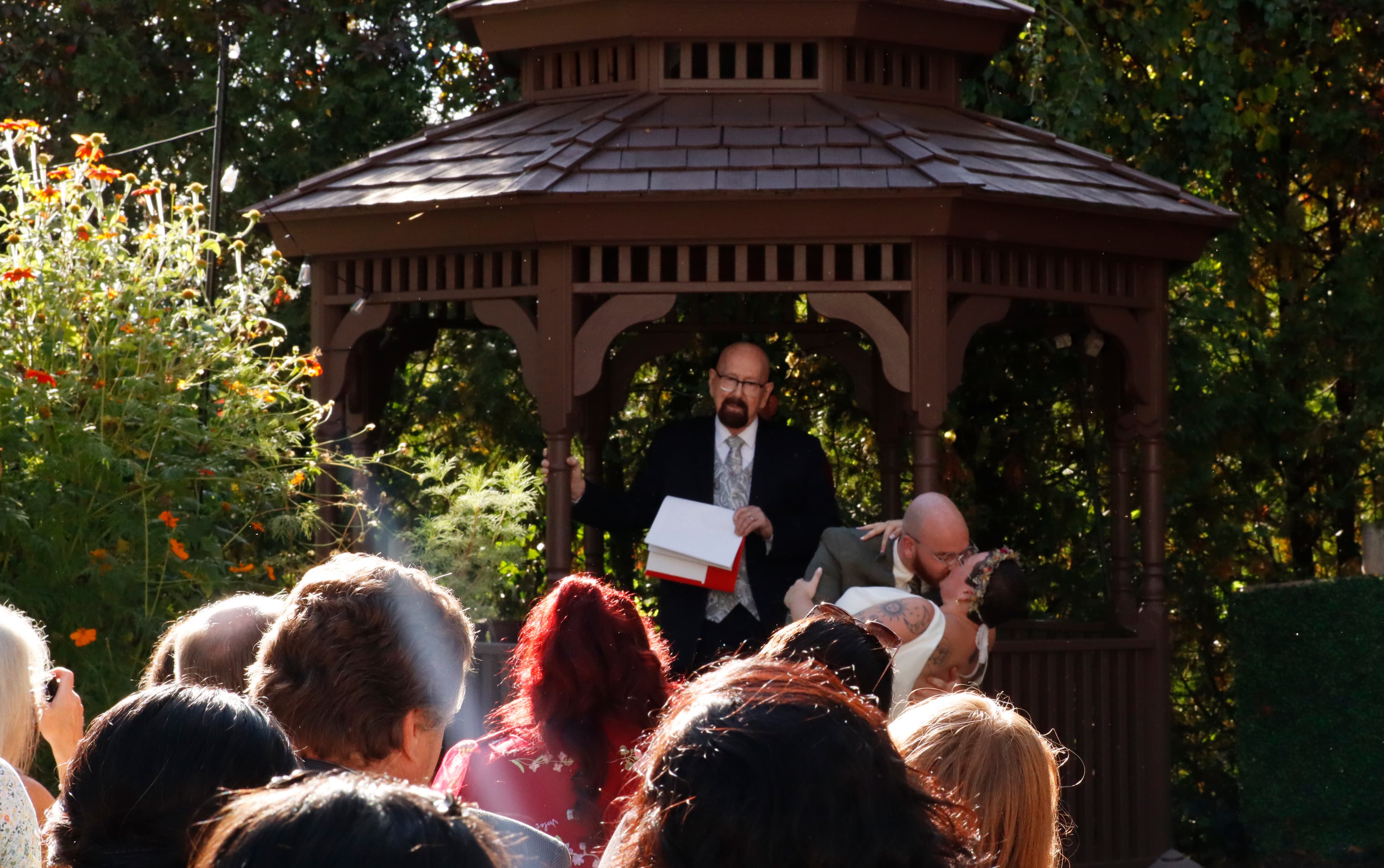 A wedding officiant stands under a gazebo addressing guests, while a couple shares a kiss.