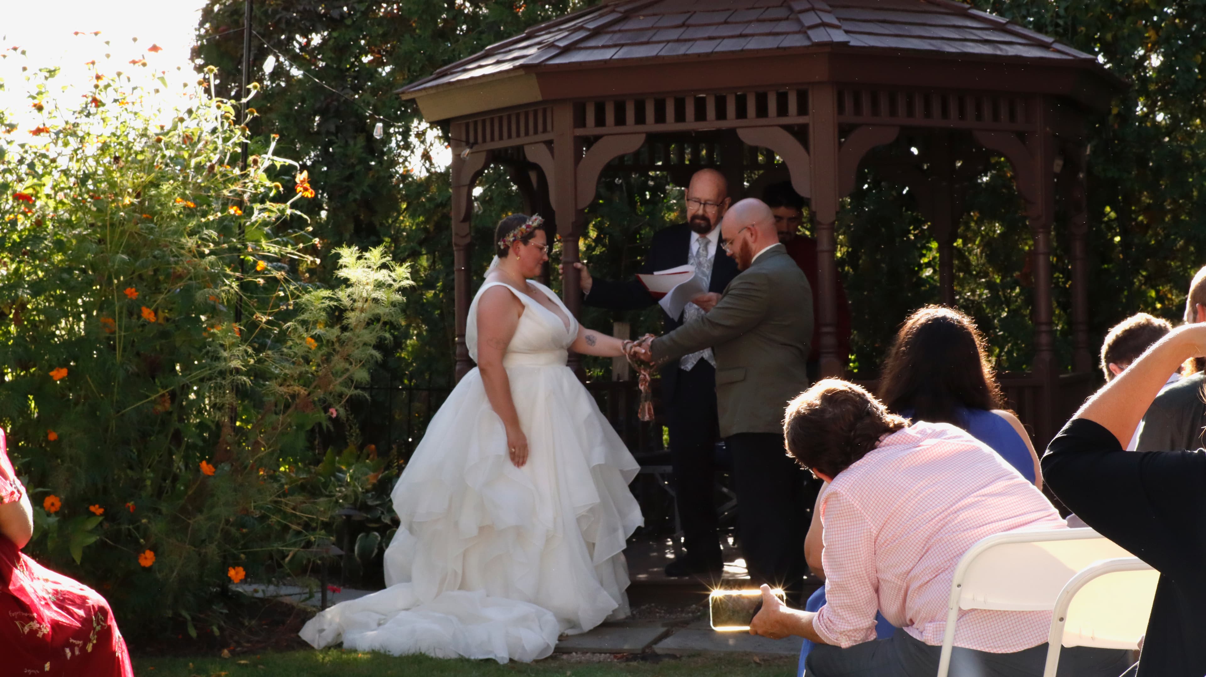 A bride and groom exchange vows under a gazebo during a wedding ceremony, with guests seated nearby.