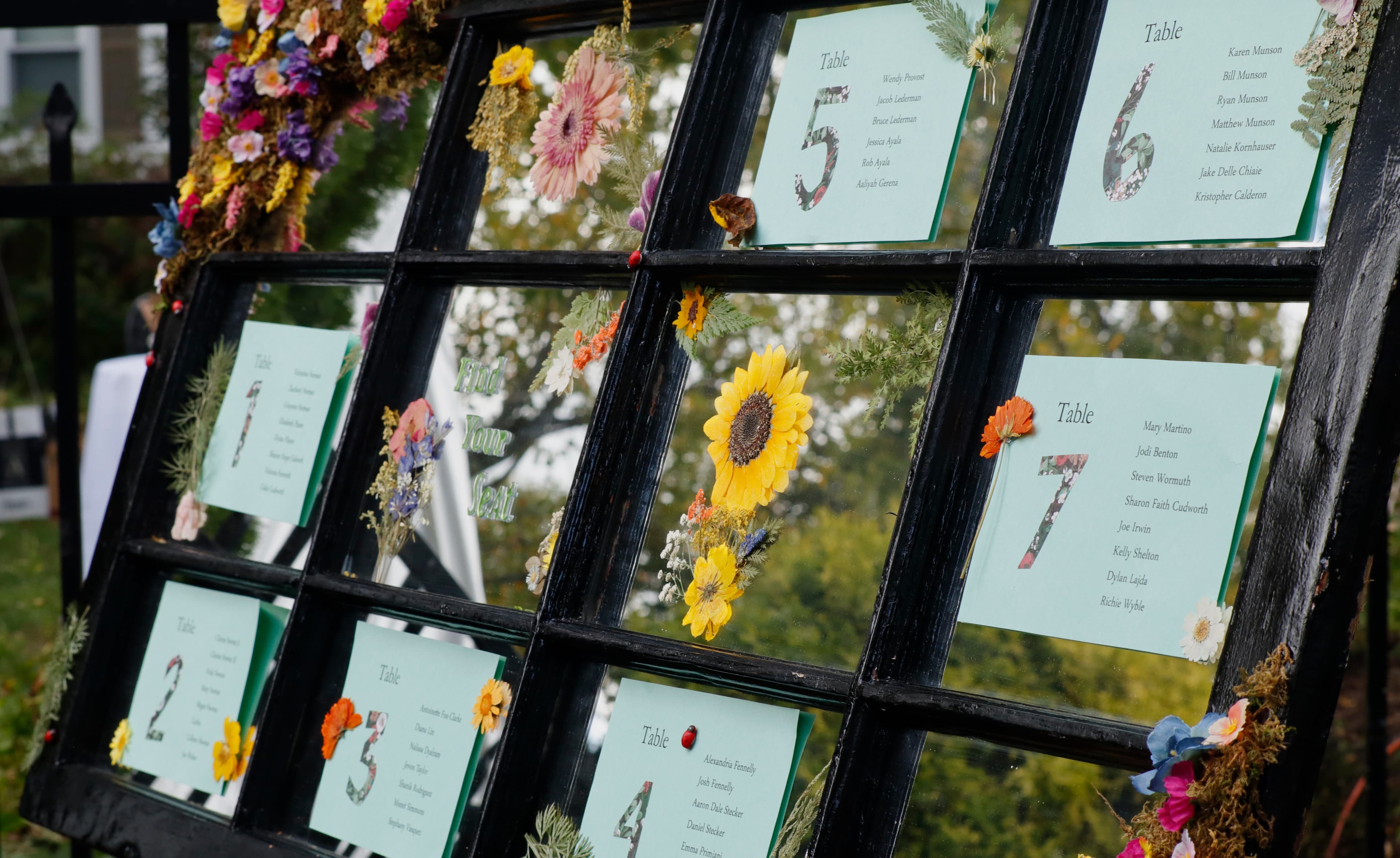 A decorative seating chart displayed on a vintage window frame, embellished with flowers and listing table assignments for an event.