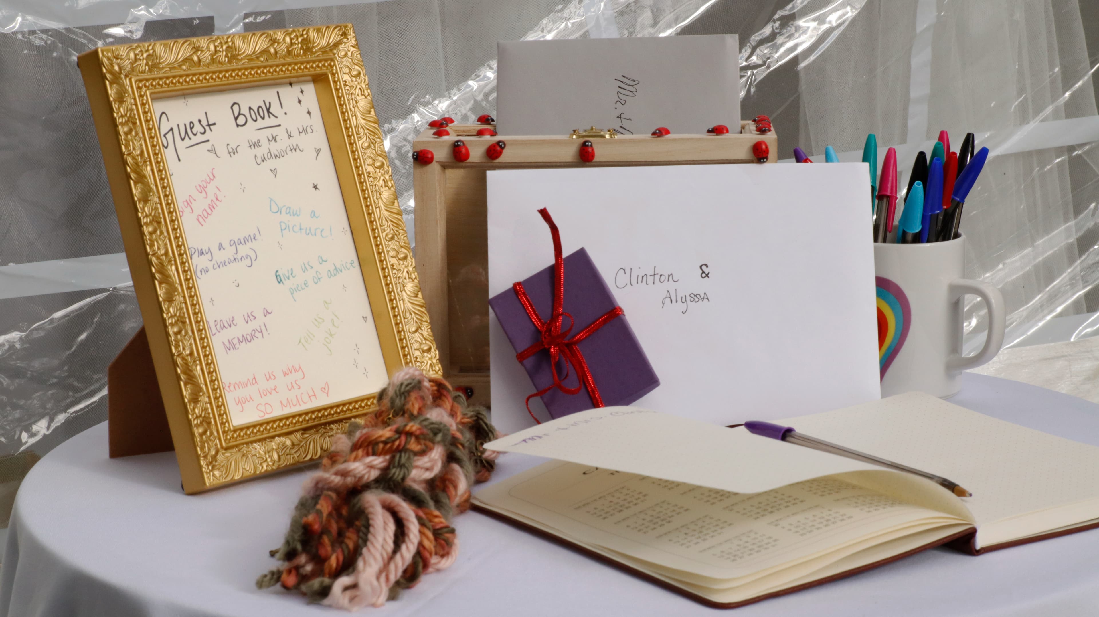 A decorative guest book setup featuring a framed sign with prompts, a notebook, colorful pens, and a small gift.