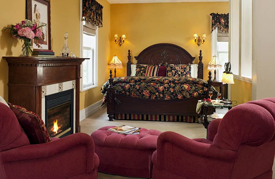 A cozy, elegantly decorated bedroom featuring a large floral-patterned bed, a fireplace, and plush seating.