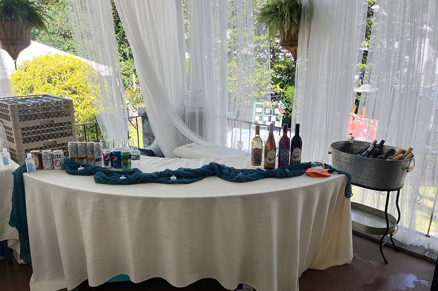 A table set for a drinks station featuring various alcoholic beverages, soft drinks, and glasses, with sheer curtains in the background.