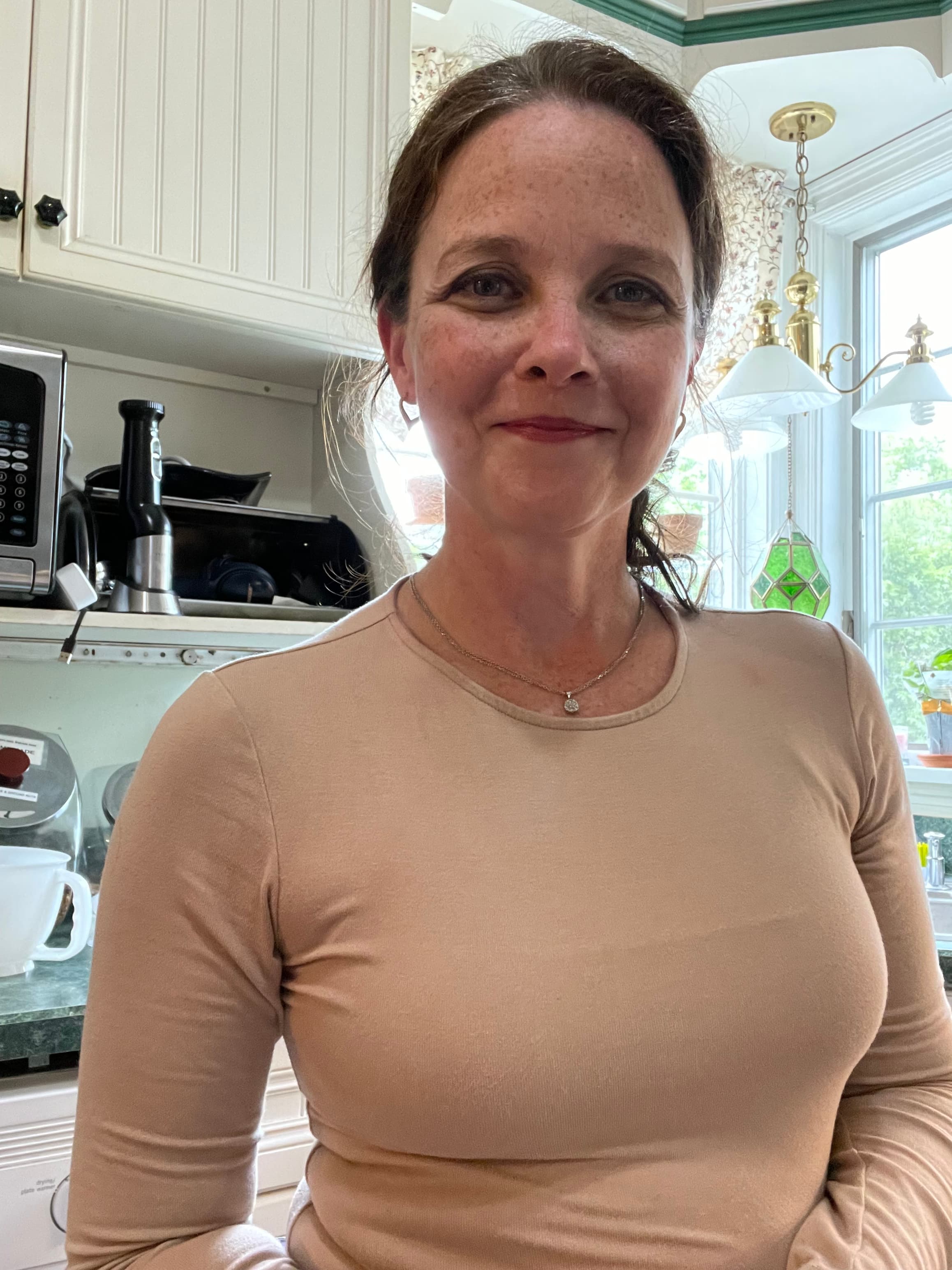 A woman with long hair stands in a bright kitchen, smiling at the camera.