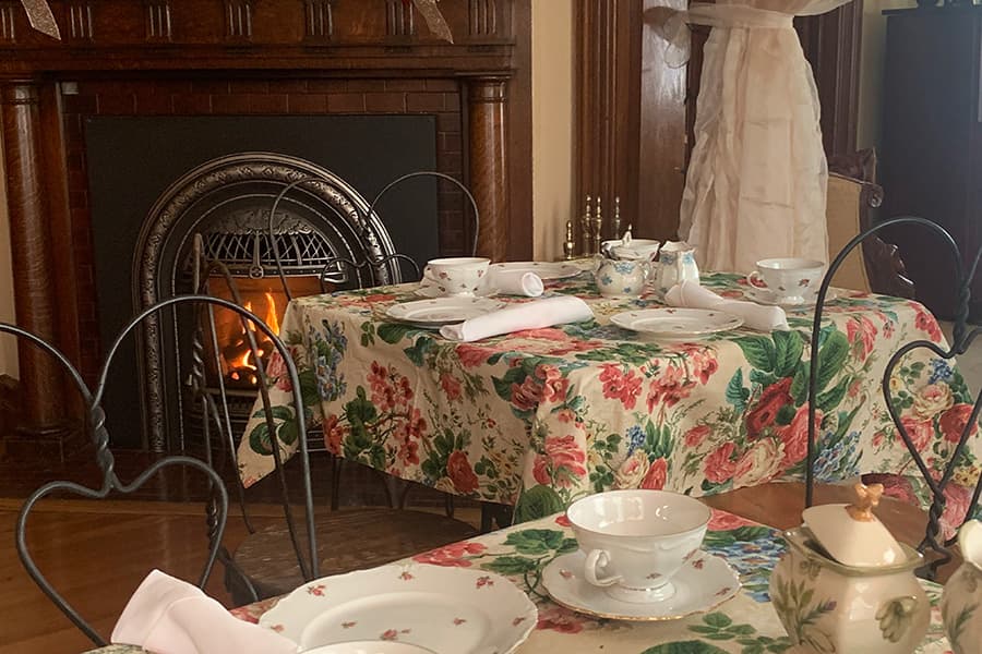 A cozy dining area features floral tablecloths, fine china, and a warm fireplace.