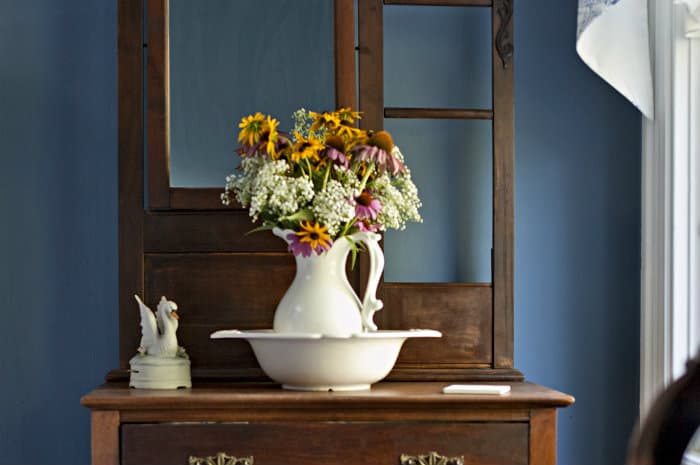 A close-up shows a wooden dresser with a white pitcher of wildflowers in a basin, set against a blue wall.