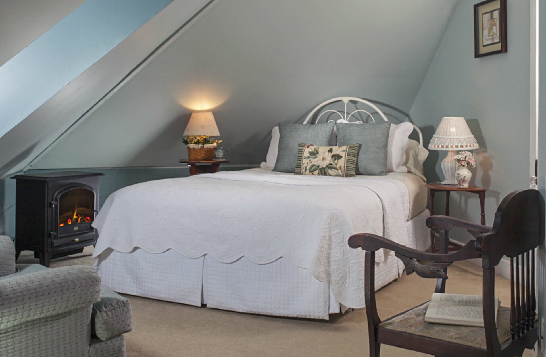 A charming attic bedroom features a white queen bed, a small fireplace, and a cozy armchair, all beneath a sloped ceiling.