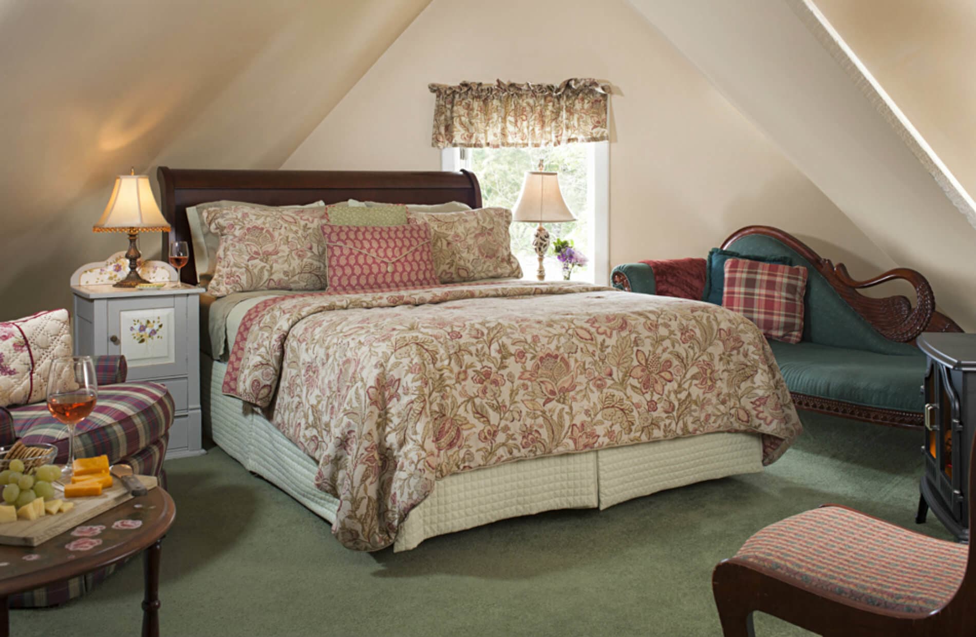 A cozy attic bedroom features a queen bed, a chaise lounge, a fireplace, and a small table with snacks, all under a sloped ceiling.