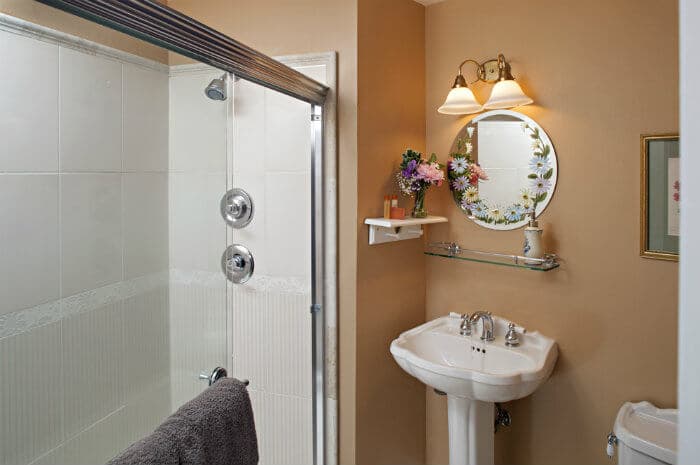 A cozy bathroom with a glass-enclosed shower, a pedestal sink, and a floral-framed mirror, illuminated by vanity lights.